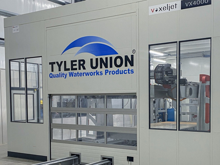 A picture of the VX4000 3D sand printer from voxeljet, installed and showing the logo of Tyler Union Quality Waterworks Products. In front of the machine three large, green and 3D printed sand molds can be seen.